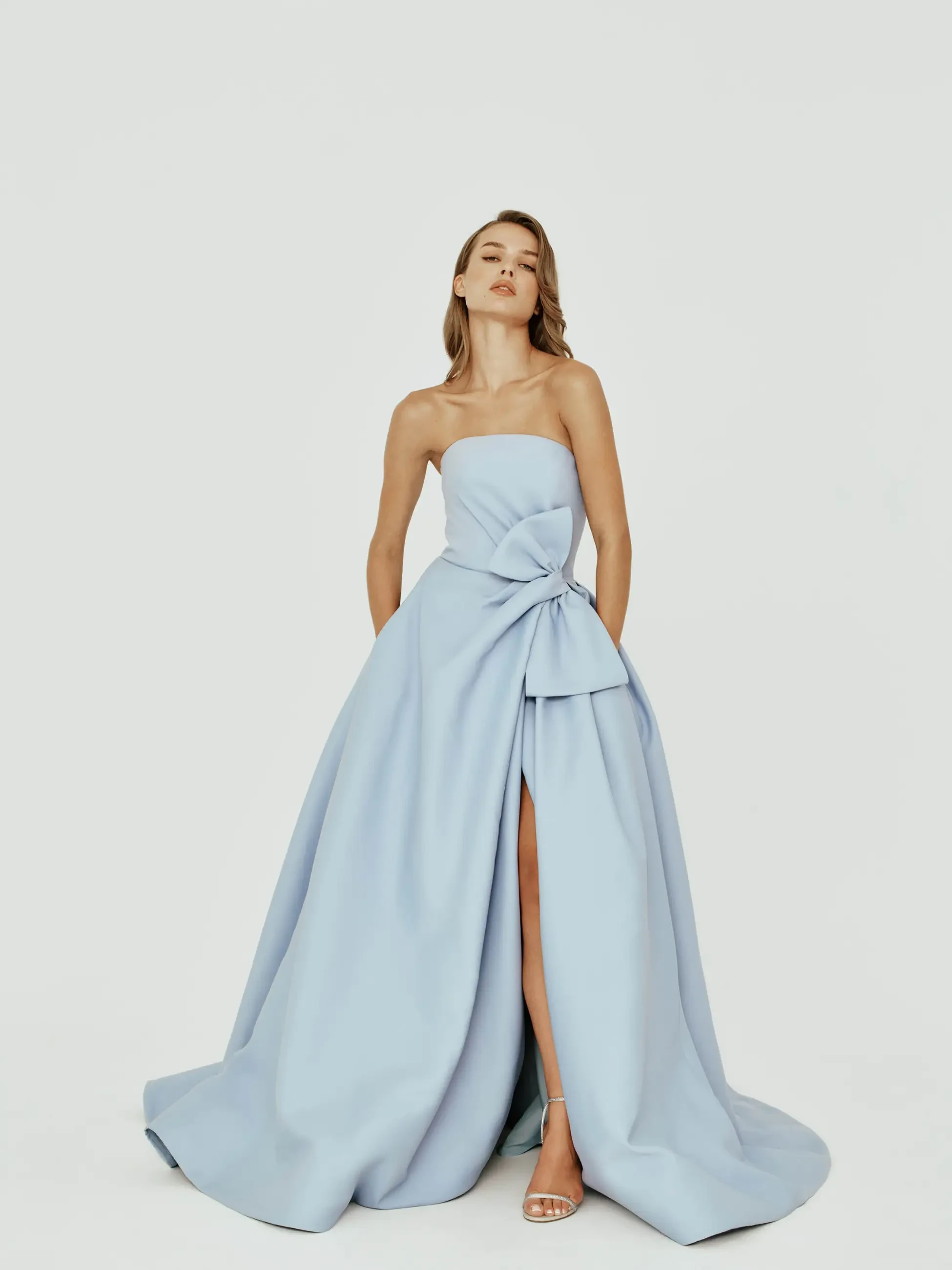 A woman poses confidently in a strapless, light blue gown with a large bow and a thigh-high slit, exuding elegance against a plain white background.